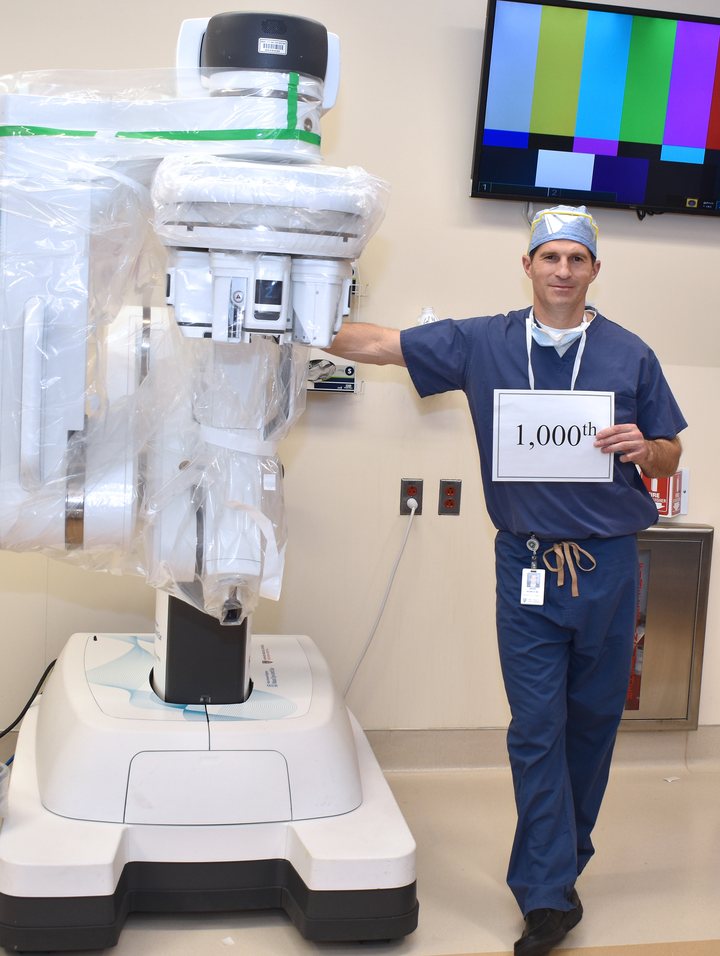 Dr. Jeremy Richmon beside the surgical robot, holding a sign reading 1,000th
